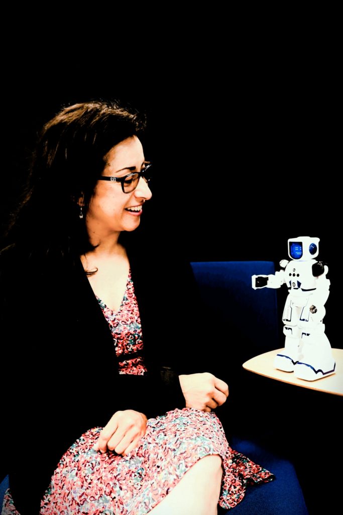 Andréa Quintero, consultante IA chez IAQ Consulting et cheffe de projet en formation e-learning spécialisée dans l’accompagnement en intelligence artificielle et la transition numérique. Andréa est assise et regarde un robot de l'intelligence artificielle